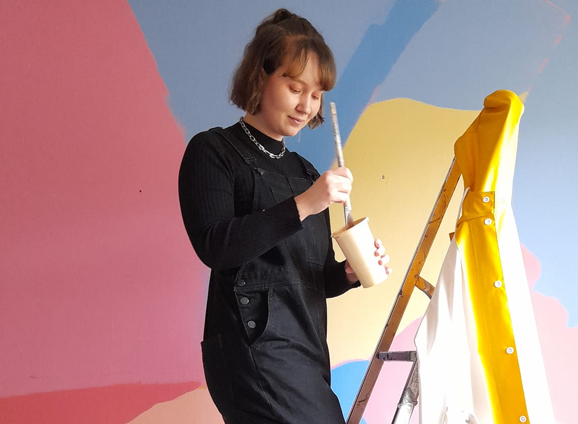 An artist stands on a ladder in front of a large mural.