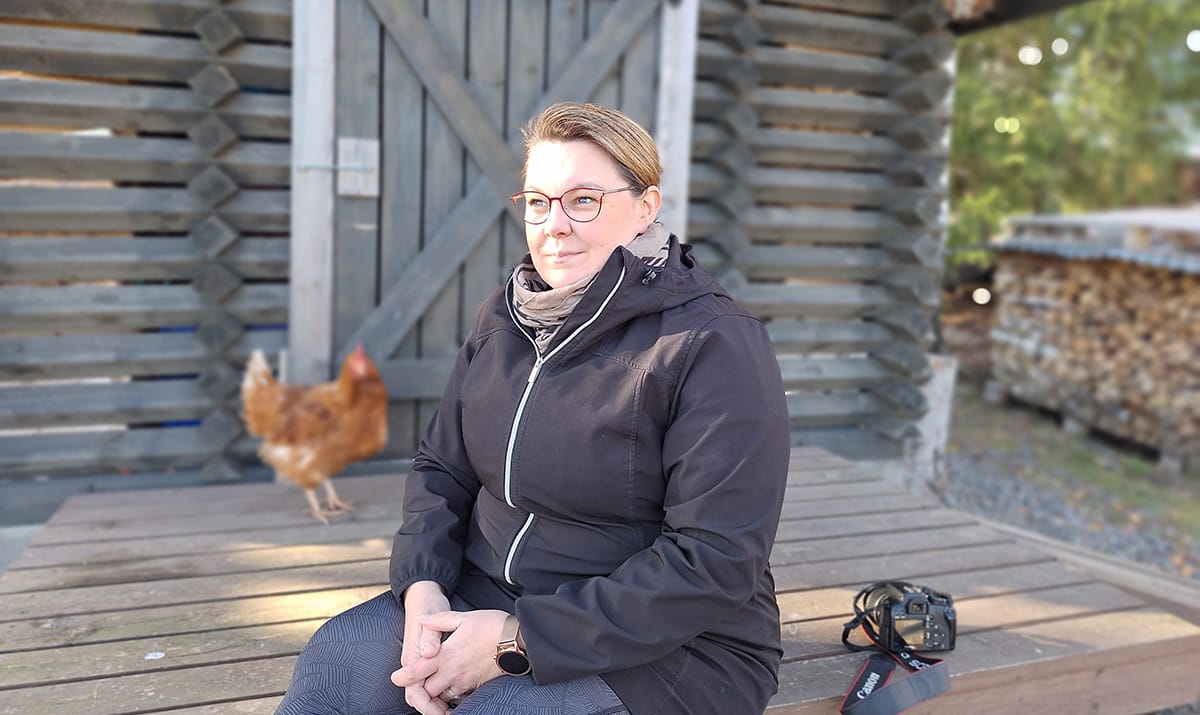 Pirita Huhtasalo istuu aitan portailla. Takana näkyy kana ja pino polttopuita.