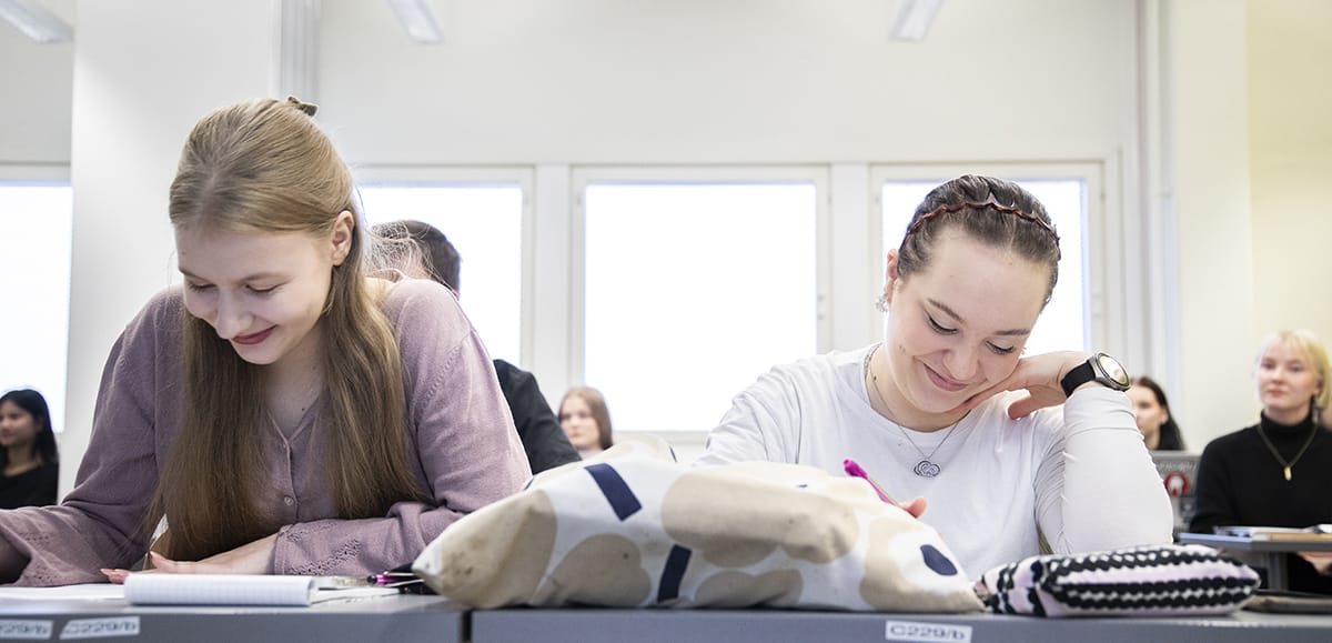 Kaksi opiskelijaa kumartuneena työpöytien ääreen. Two students bent over their desks.