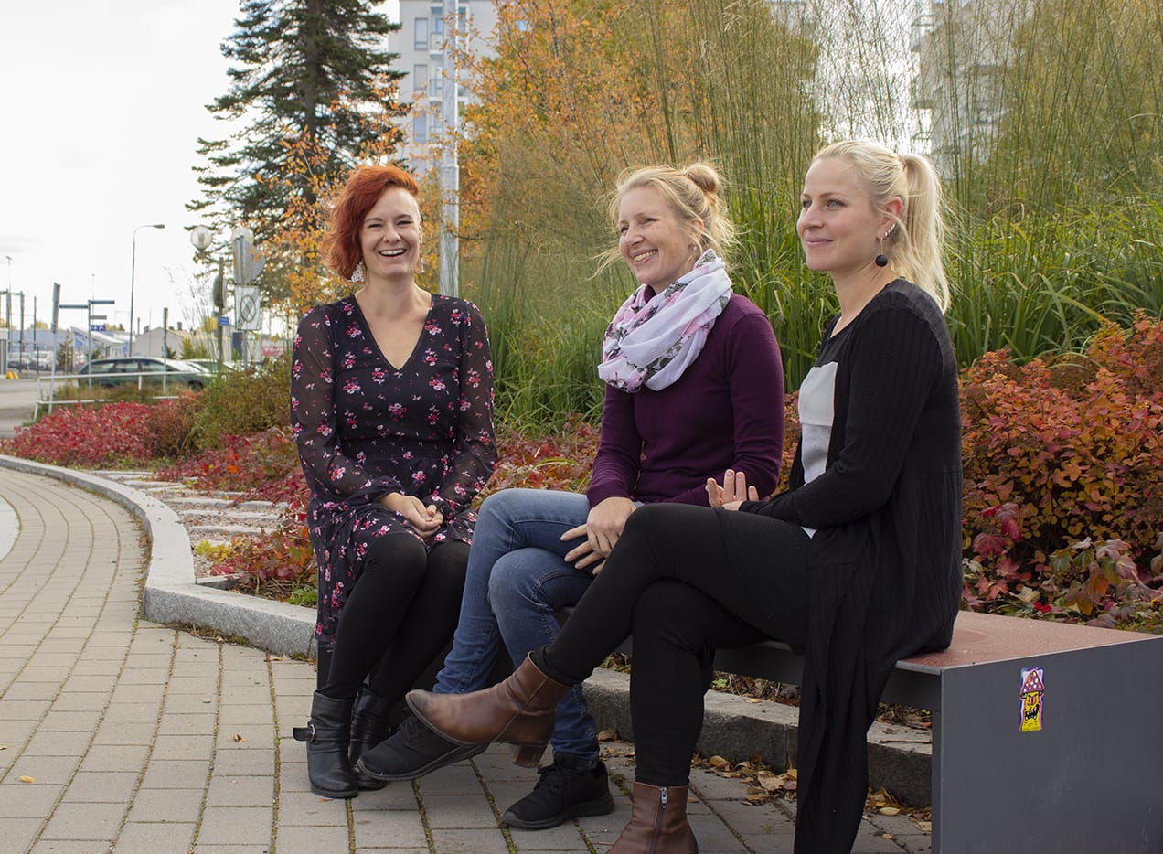 Kolme naista istuu penkillä, taustalla ruskan värejä.