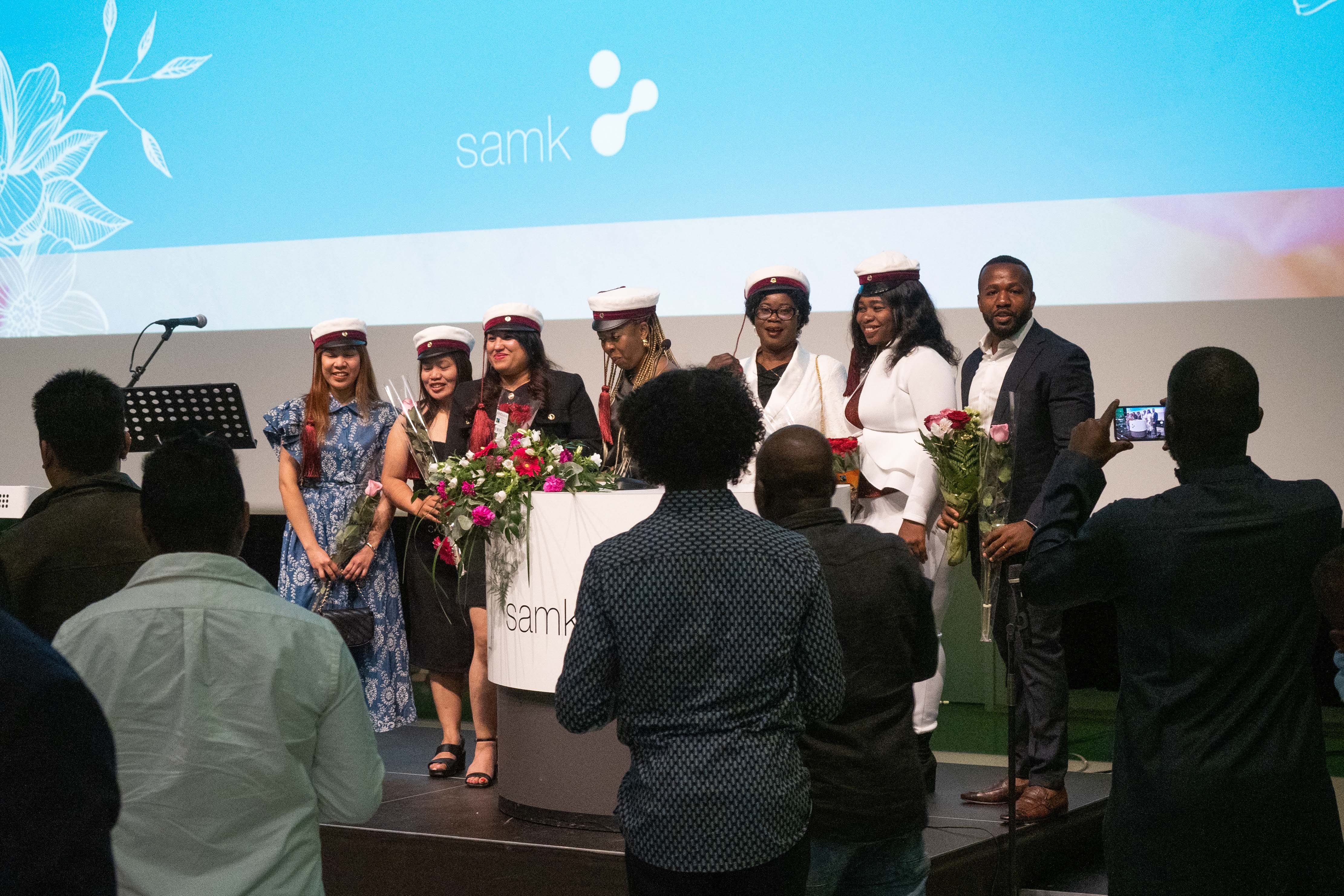 Ryhmä valmistuvia opiskelijoita seisoo lavalla SAMK-screenin edessä.