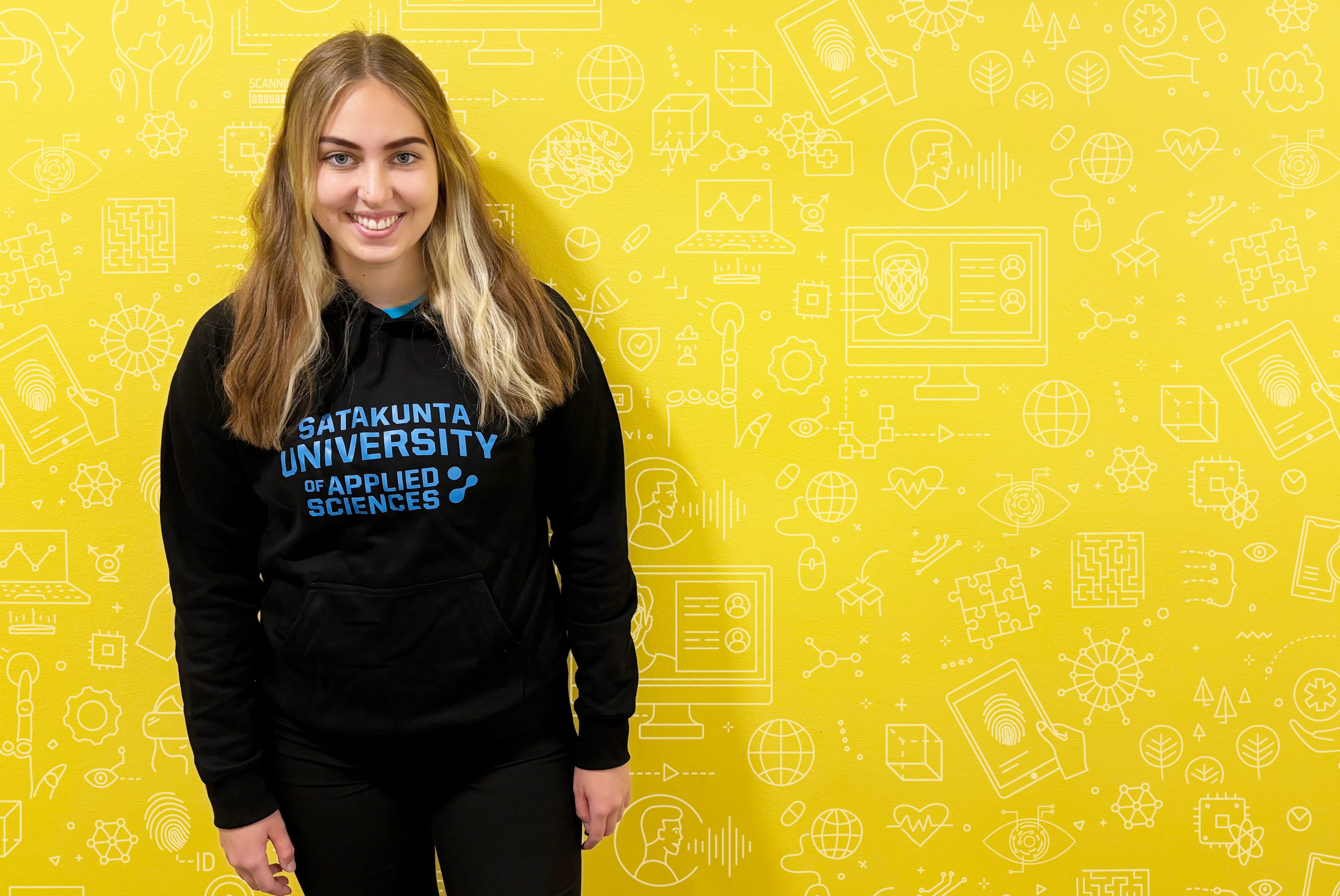 A student standing in front of a yellow wall wearing a SAMK hoodie.